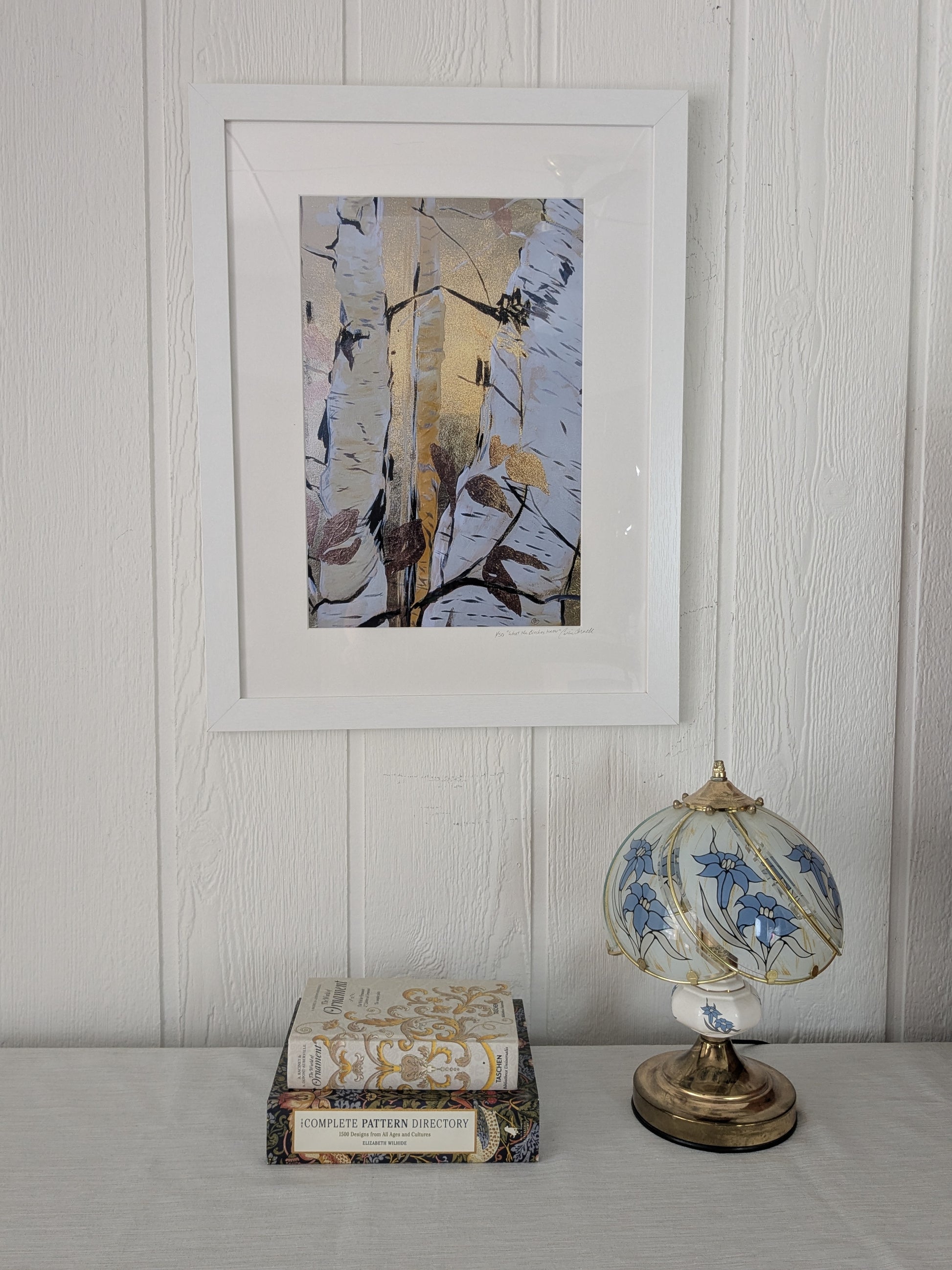 A framed abstract art print with white, beige, and brown brushstrokes hangs on a white paneled wall above a table with a vintage patterned book and a blue-and-white patterned table lamp.